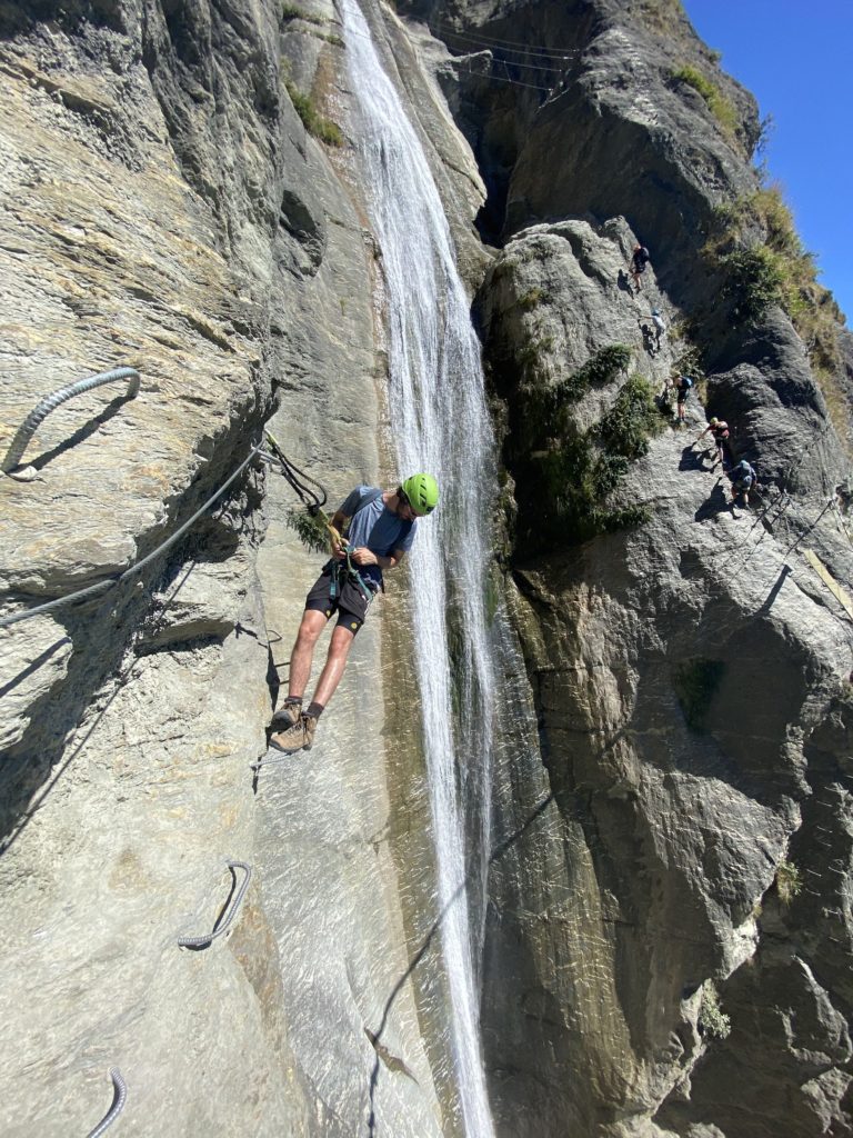 ben hangs over edge of via ferrata with waterfall in the background