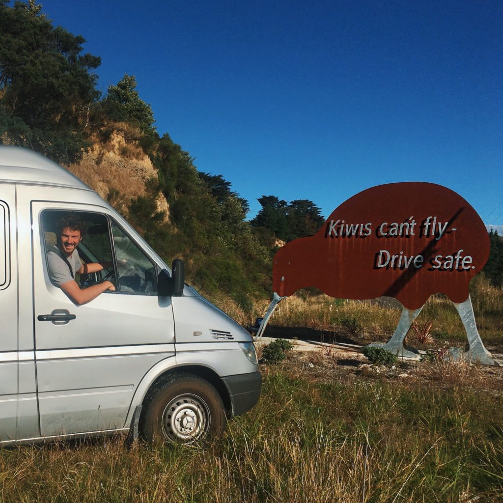 how to plan a new zealand road trip: ben in the van with a sign in the shape of a kiwi that says "kiwis can't fly -- drive safe!"