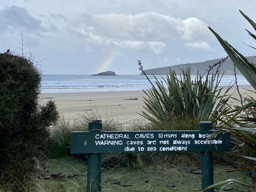 Cathedral Caves sign with beach and rainbow in the background