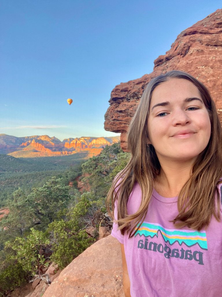 Niki takes a selfie in front of a hot air balloon and mountains at sunrise on the Devil's Bridge hike, Sedona, Arizona