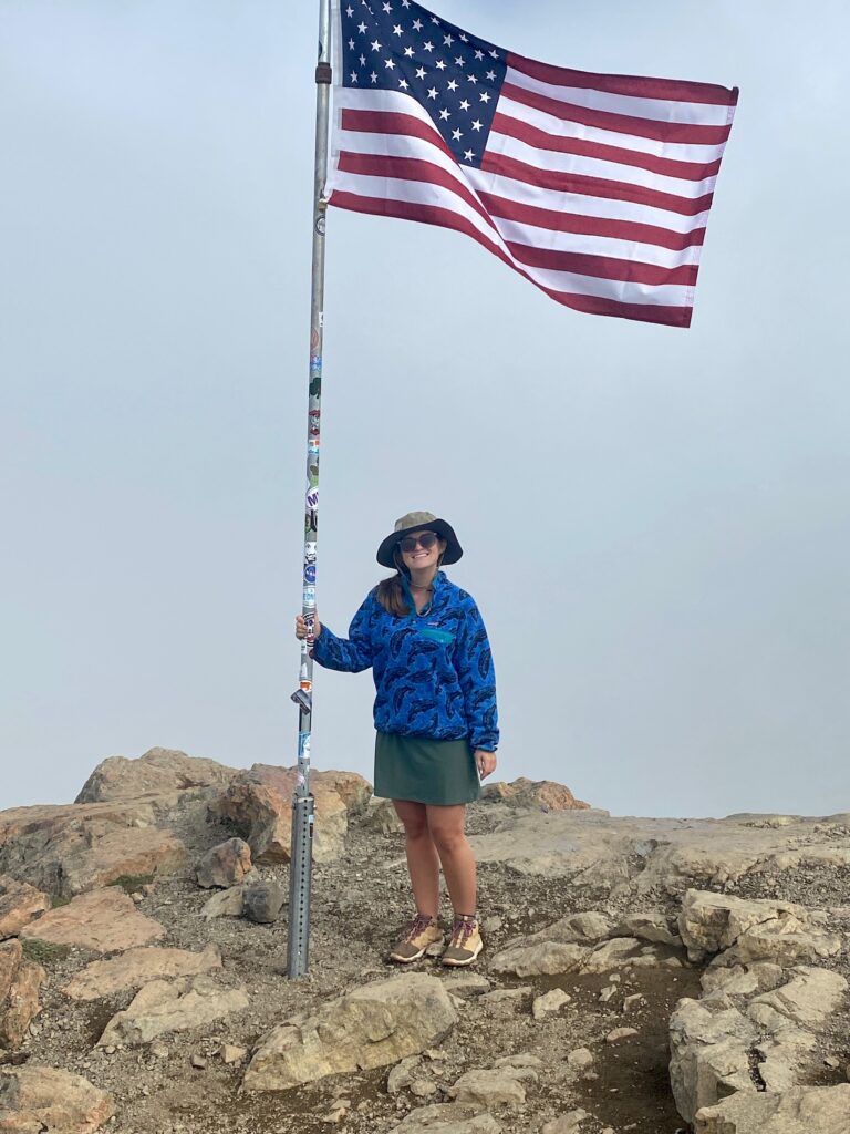 Niki stands in front of an American flag at the mountain peak