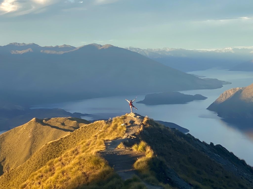 Niki stands at Roys Peak viewpoint at sunset with Lake Wanaka in the background