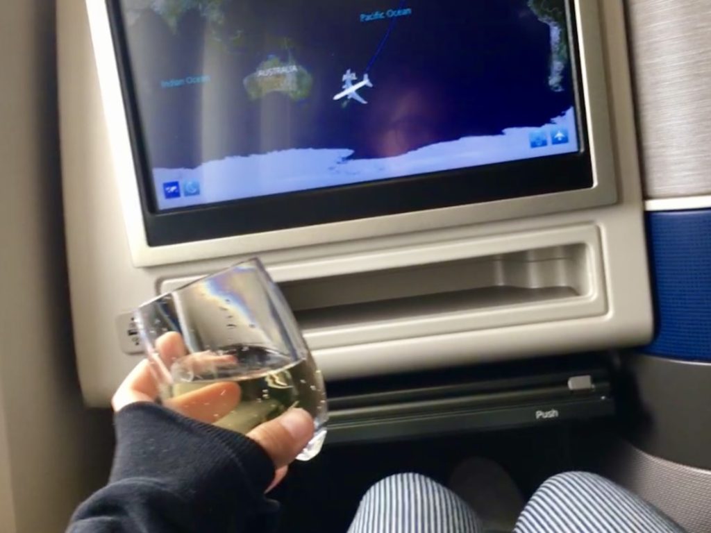 Niki holds a glass of champagne in front of her airplane seat back screen in Auckland, New Zealand