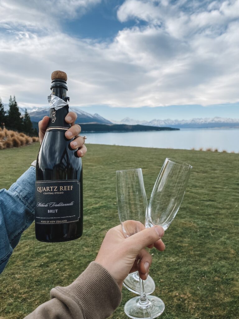 Niki and Ben hold a bottle of Quartz Reef prosecco and two glasses with a view of Lake Pukaki and Aoraki/Mt Cook in the distance, South Island, New Zealand