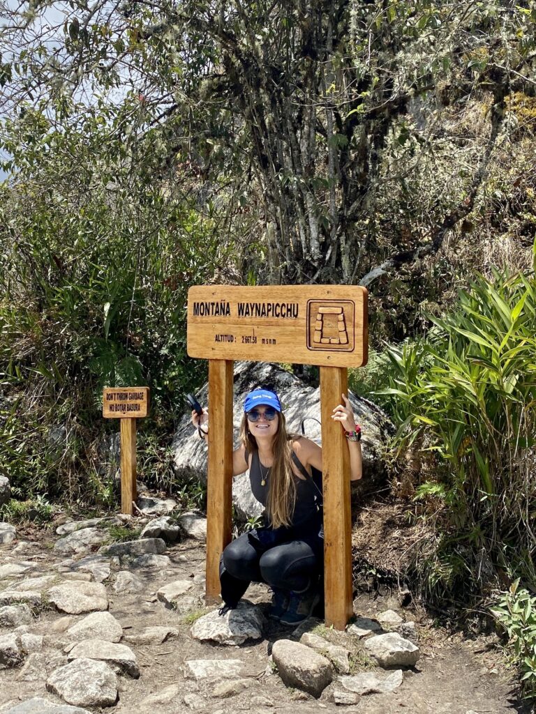 Niki sits under the Wayna Picchu sign while hiking Huayna Picchu, Peru